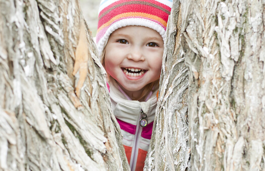 A child wearing a beanie smiles through the fork in a tree trunk