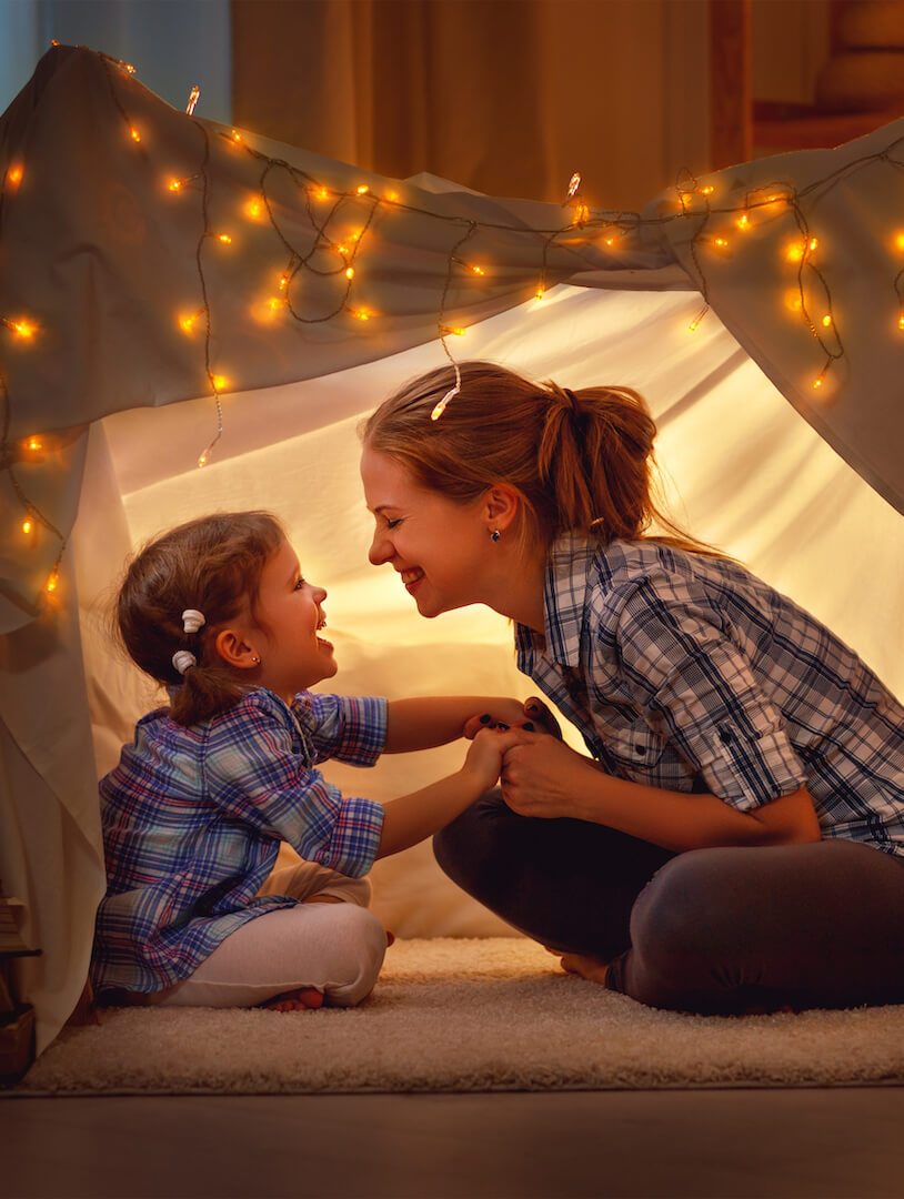 Woman holds little boys hands. They sit cross legged in a fairylit tent